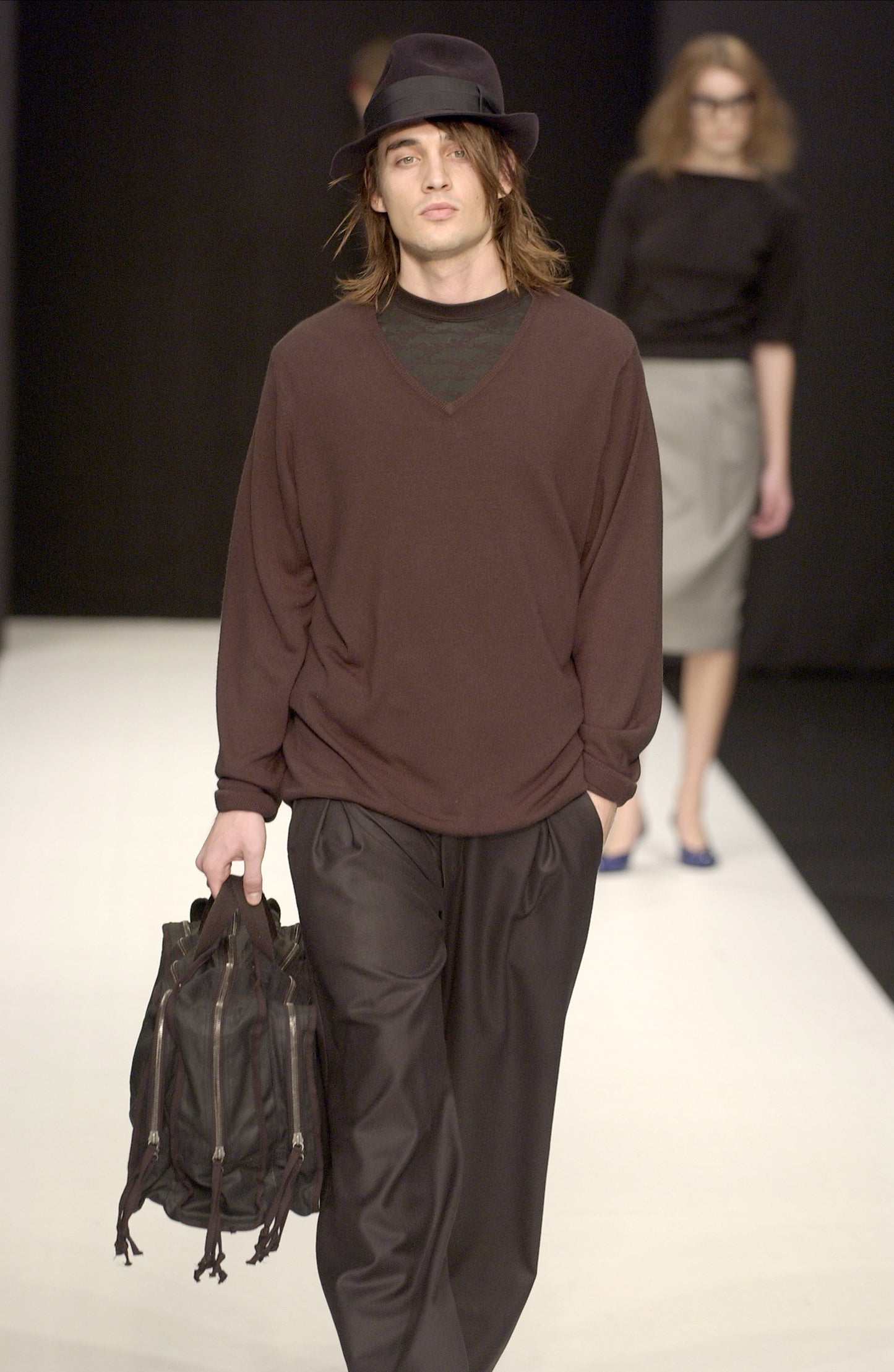 Man walking on a runway wearing a STÆRK brown and black outfit with a hat and carrying a bag.