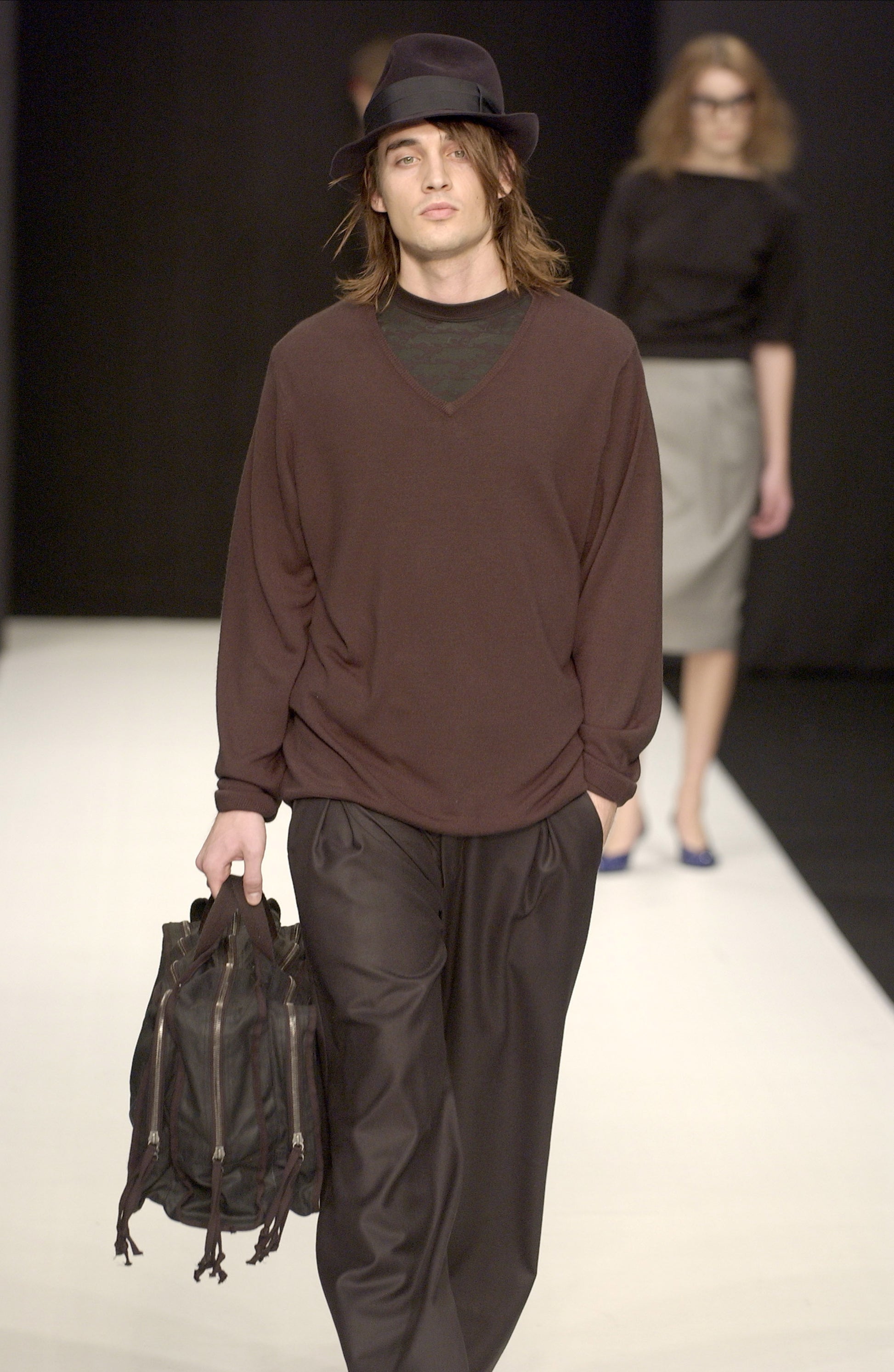 Man walking on a runway wearing a STÆRK brown and black outfit with a hat and carrying a bag.